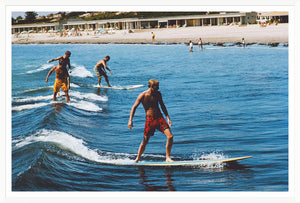 Four people surf small waves near a sandy beach, sunbathers and buildings behind them—a scene from "Surfing Brothers" by Slim Aarons (Getty Images, 1965), available from Soicher Marin.