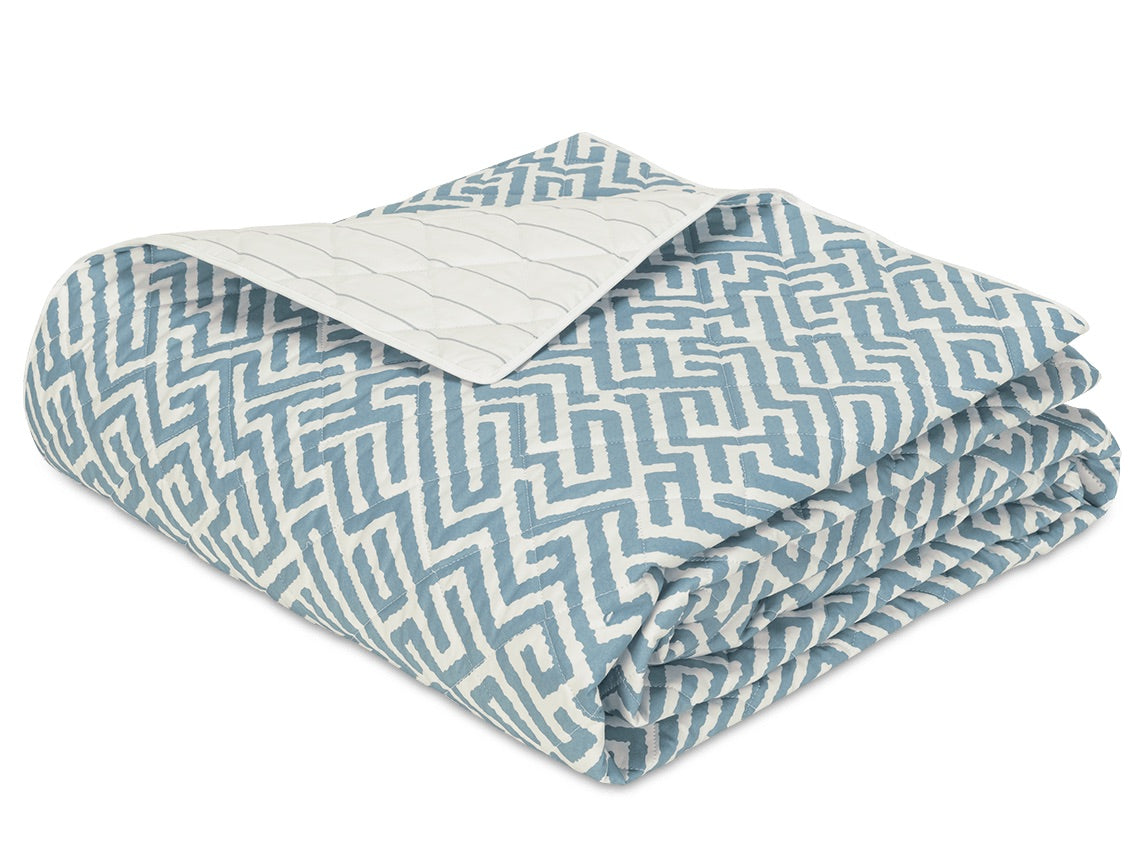 The Gabriel Bedding Collection by Matouk showcases a folded quilt with a blue and white geometric motif, partially turned to display its solid white quilted underside.