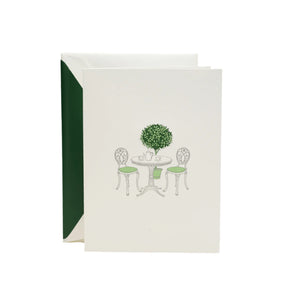 The Crane Garden Bistro Card Set features pearl white folded notes engraved with a bistro table, chairs, and a potted topiary, complemented by green-lined envelopes—perfect for sophisticated correspondence.