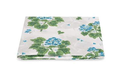 A folded white fabric with a green leafy and blue floral print from the Tillett x Matouk Geranium Bedding Collection, Blue by Matouk, placed against a plain white background.