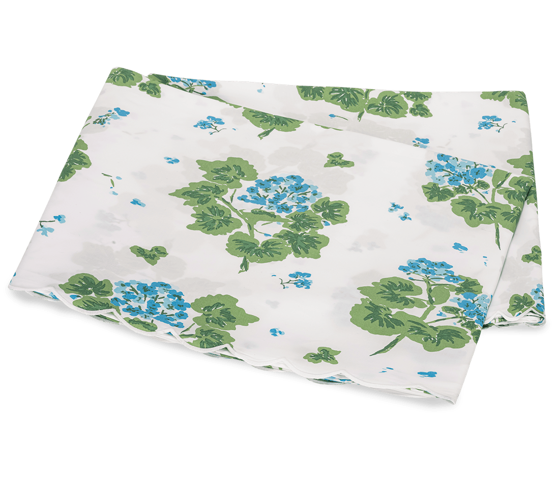 The Tillett x Matouk Geranium Bedding Collection in Blue by Matouk showcases folded white percale fabric with a blue and green floral pattern and scalloped edges, displayed against a white background.
