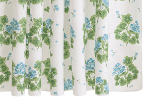 The Matouk Geranium Shower Curtain Collection features a white curtain adorned with green leaves and small blue flowers, inspired by the classic Geranium print and crafted in Luca percale style.