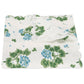 The Matouk Tillett x Matouk Blue Geranium Table Linen Collection features a folded white linen tablecloth with blue and green floral design and scalloped edges, perfect for elegant tables.