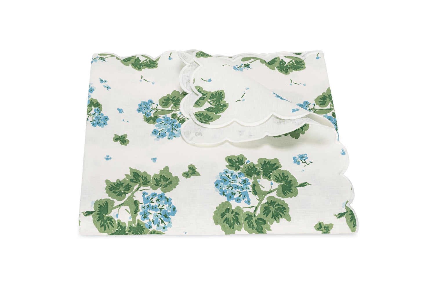 The Matouk Tillett x Matouk Blue Geranium Table Linen Collection features a folded white linen tablecloth with blue and green floral design and scalloped edges, perfect for elegant tables.