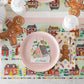 A gingerbread-themed Gingerbread Partyware Collection placemat and napkin set the table, featuring a pink plate with a house-shaped name card, coordinating decor, and mugs of hot cocoa.