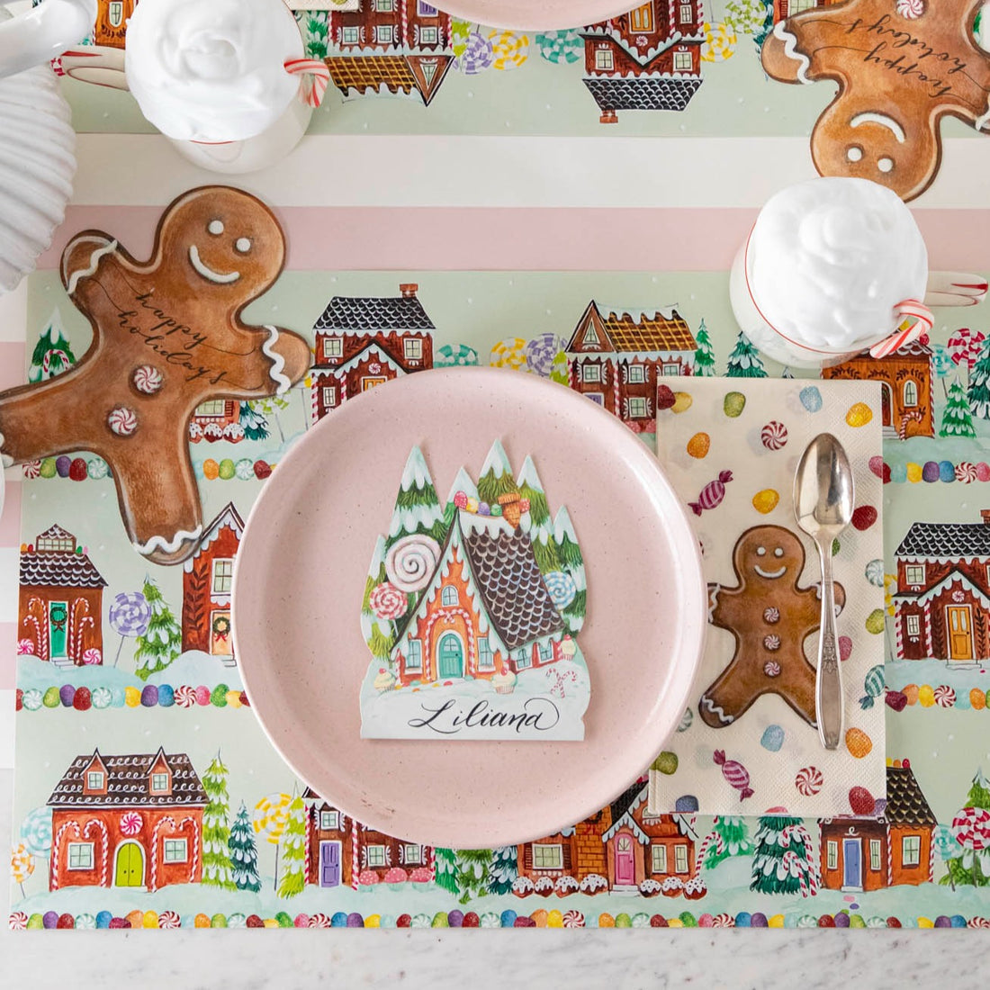 A gingerbread-themed Gingerbread Partyware Collection placemat and napkin set the table, featuring a pink plate with a house-shaped name card, coordinating decor, and mugs of hot cocoa.