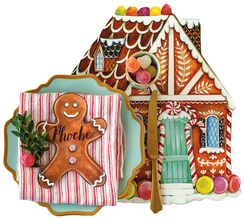 A Gingerbread Partyware Collection placemat shaped like a gingerbread house with a blue plate, red-striped napkin, “Phoebe” gingerbread man place card, gold flatware, and gumdrops.