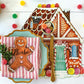 A gingerbread house-shaped placemat from the Hester & Cook Gingerbread Partyware Collection, set with gold cutlery, gumdrops, and a gingerbread man name card on a striped napkin atop a white table.