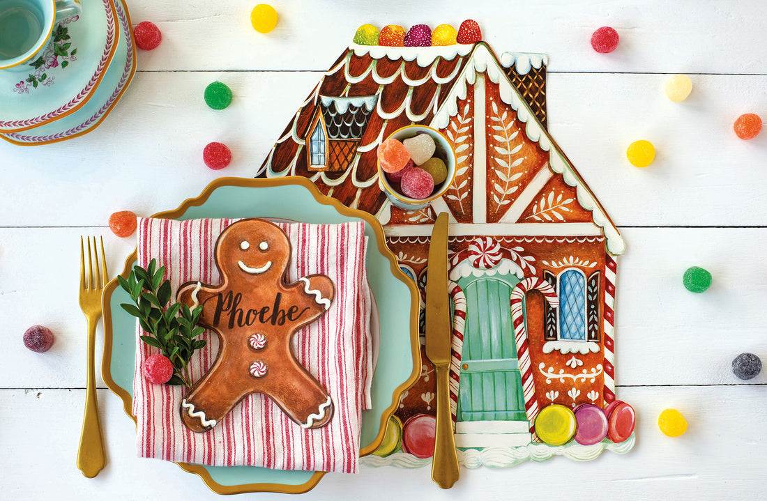 A gingerbread house-shaped placemat from the Hester & Cook Gingerbread Partyware Collection, set with gold cutlery, gumdrops, and a gingerbread man name card on a striped napkin atop a white table.