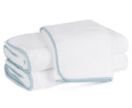 Three neatly folded white towels from the Matouk Giza Bath Collection, featuring light blue edging and crafted from luxurious Giza cotton, are stacked on a white surface.