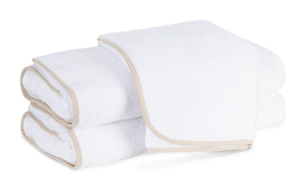 Two white towels with beige trim from the Matouk Giza Bath Collection, Sand, are neatly folded. Crafted from luxurious Giza cotton, one towel is partially unfolded on top.