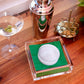 A cocktail setup features a martini, metal shaker, gold holder, and the Tara Wilson Designs Lucite Cocktail Napkin Holder with Golf Ball Weight and green napkins—an ideal cocktail hour accessory on a wooden surface.