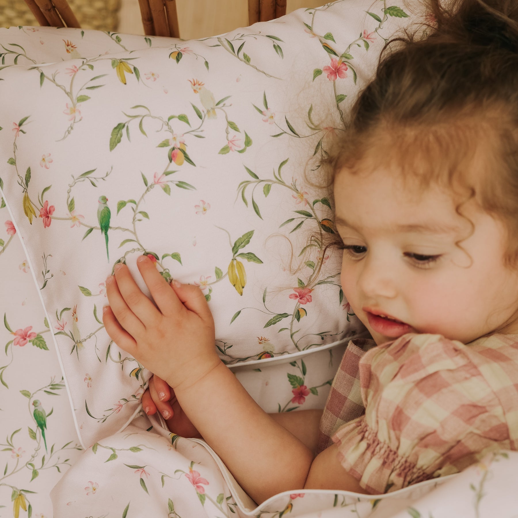 A young child in a plaid dress gently touches the Gooselings Dreaming of Paradise Toddler Pillow, its floral pattern perfect for comforting little sleepers and inspiring sweet dreams.