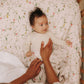 A baby lies on the Gooselings Dreaming of Paradise Crib Sheet, featuring a floral pattern and OEKO-TEX certification, while an adult gently adjusts the blanket.