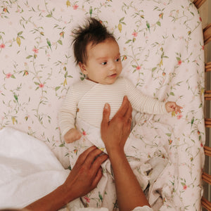 A baby lies on the Gooselings Dreaming of Paradise Crib Sheet, featuring a floral pattern and OEKO-TEX certification, while an adult gently adjusts the blanket.