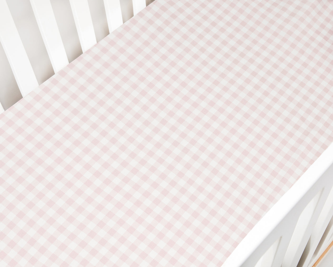 A close-up of a Gooselings Picnic Gingham Pink Crib Sheet, made from extra soft cotton sateen in pink and white, covering a crib mattress inside a white crib.