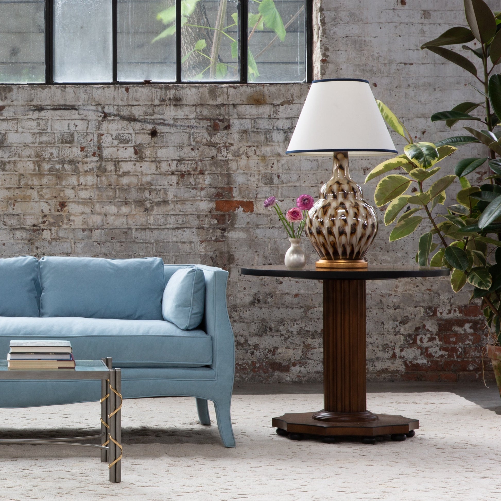 A light blue sofa sits next to the Bunny Williams Gottlieb Side Table, topped with a lamp and vase of flowers, set against a brick wall with large windows and potted plants.