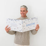 A man in a beige sweater smiles as he holds and reads the large “Snow” book, featuring an illustrated skiing cover, capturing the excitement of the Snow Book Signing with Gray Malin by Hive Events at West Palm Beach.