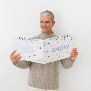 A man in a beige sweater smiles as he holds and reads the large “Snow” book, featuring an illustrated skiing cover, capturing the excitement of the Snow Book Signing with Gray Malin by Hive Events at West Palm Beach.