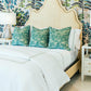 The Ingrid Bed by Oly features a beige raffia-upholstered headboard, complemented by three green pillows, white bedding, and matching white nightstands with lamps, all set against tropical-themed wallpaper.
