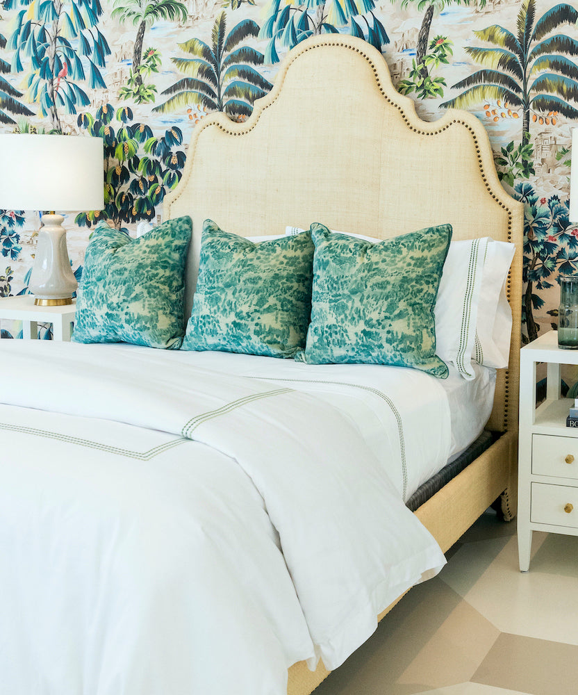 The Ingrid Bed by Oly features a beige raffia-upholstered headboard, complemented by three green pillows, white bedding, and matching white nightstands with lamps, all set against tropical-themed wallpaper.