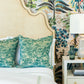 The Ingrid Bed by Oly features a beige upholstered headboard with nailhead detailing, paired with two green patterned pillows, white bedding, and a nightstand with lamp set against tropical wallpaper.