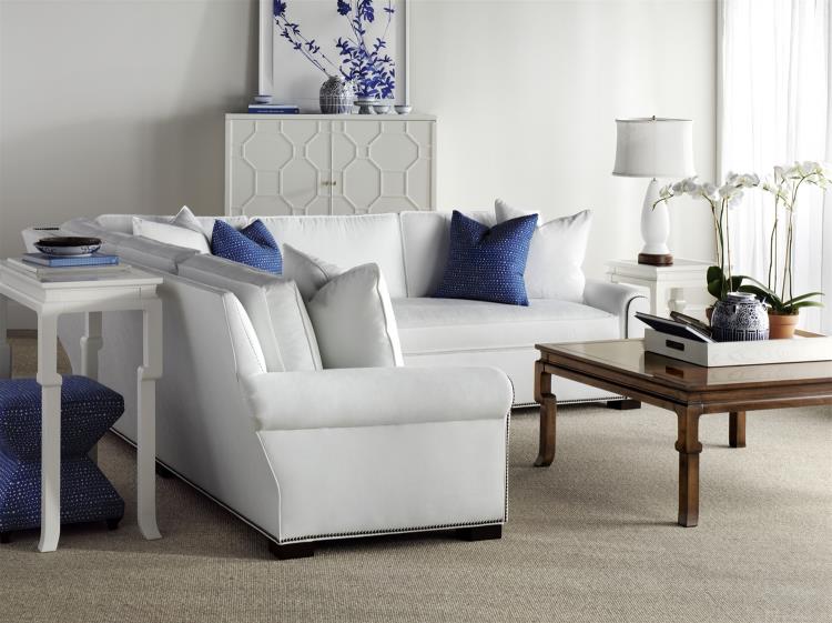 A modern living room with white furniture, blue accent pillows, a Hickory Chair Ceylon Cocktail Table, a decorative cabinet, and a table lamp beside a window with sheer curtains brings the sophisticated charm of West Palm Beach into your home.