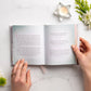 Hands cradle an open book surrounded by flowers and a lit candle on a light surface, creating a serene scene ideal for devotions, featuring "Jesus Calling For Moms" by Thomas Nelson.