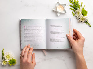 Hands cradle an open book surrounded by flowers and a lit candle on a light surface, creating a serene scene ideal for devotions, featuring "Jesus Calling For Moms" by Thomas Nelson.