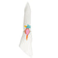 A white cloth napkin is rolled in a ring and accented with an embroidered colorful bird design in blue, pink, orange, and yellow thread. The napkin is held by the Mercedes Salazar Cockatoo Napkin Ring, handmade in Colombia from natural raffia fiber.