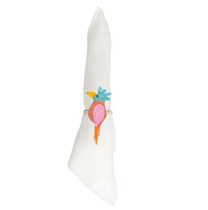 A white cloth napkin is rolled in a ring and accented with an embroidered colorful bird design in blue, pink, orange, and yellow thread. The napkin is held by the Mercedes Salazar Cockatoo Napkin Ring, handmade in Colombia from natural raffia fiber.