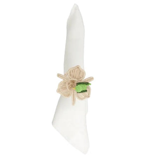 A white cloth napkin is folded and held by the Mercedes Salazar Pajarito Napkin Ring, handmade in Colombia from raffia fiber, featuring a beige floral design with a small green accent in the center.