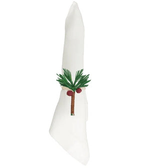 A white cloth napkin folded upright with an embroidered green palm tree and two coconuts on the front, elegantly accented by the Mercedes Salazar Coconut Palm Tree Napkin Ring, handmade in Colombia.