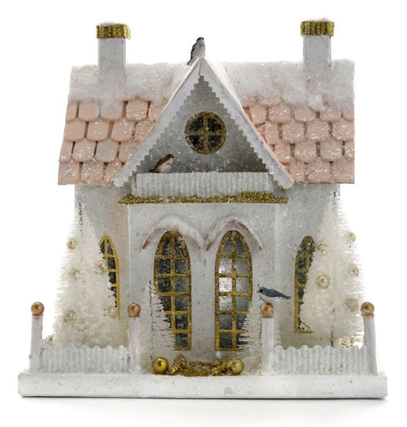 The Cody Foster Winter Cottage by Cody Foster is a white, snowy miniature house with gold accents, two chimneys, arched windows, and decorative trees adorning the front. Crafted from cardboard.