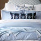 A neatly made bed features blue and white stonewashed cotton bedding, patterned pillows, a dark blue accent pillow, and the John Robshaw Daasu Throw Collection's textured light blue blanket.