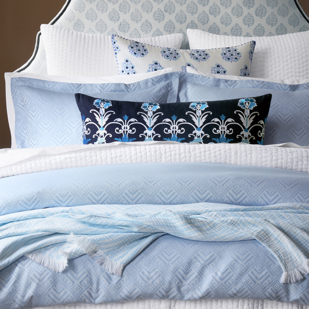 A neatly made bed features blue and white stonewashed cotton bedding, patterned pillows, a dark blue accent pillow, and the John Robshaw Daasu Throw Collection's textured light blue blanket.