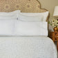 A neatly made bed with John Robshaw's Stitched Light Indigo Sheet Set, a patterned headboard, and a bedside table featuring a lamp and a vase of flowers.