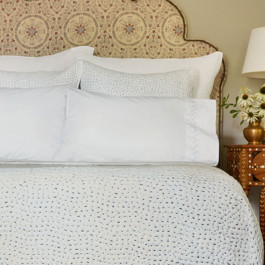 A neatly made bed with John Robshaw's Stitched Light Indigo Sheet Set, a patterned headboard, and a bedside table featuring a lamp and a vase of flowers.