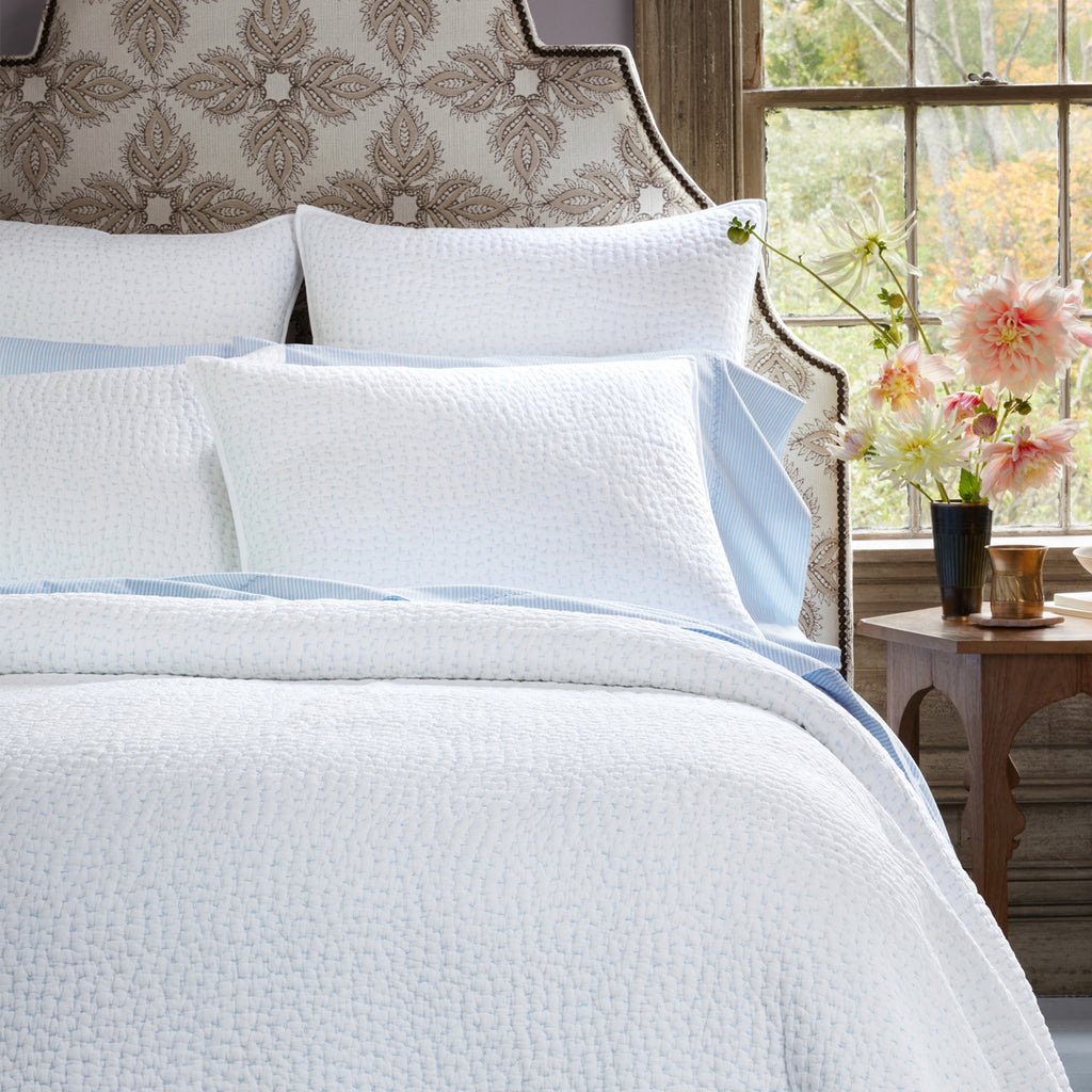 A neatly made bed featuring the John Robshaw Stitched Mist Coverlet, white textured organic cotton voile pillows, an upholstered headboard, and a bedside table with flowers and a cup by the window.