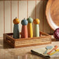 Four colorful pepper mills in the Ellie Cane Tray by Be Home sit on a kitchen counter next to a round wooden bowl, a red cabbage, and folded towels with utensils.