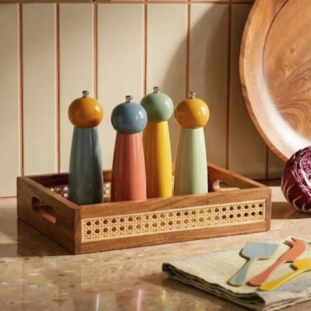 Four colorful pepper mills in the Ellie Cane Tray by Be Home sit on a kitchen counter next to a round wooden bowl, a red cabbage, and folded towels with utensils.