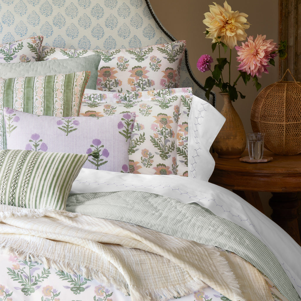 A bed styled with floral and striped pillows, pastel bedding, and a John Robshaw Daasu Throw Collection throw is set beside a round wooden nightstand featuring flowers, a glass of water, and a woven lamp.