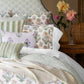 A neatly made bed features the John Robshaw Nandi Sage Coverlet, block printed and striped pillows, a patterned headboard, a throw blanket, plus a bedside table with dahlias in a vase, a wicker lamp, and a glass of water.
