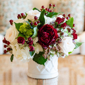 The Live Classic Holiday Floral Arrangement by Hive Floral Studio features red and white roses, hydrangeas, greenery, and red berries in a white ceramic cachepot—perfect as a festive centerpiece.