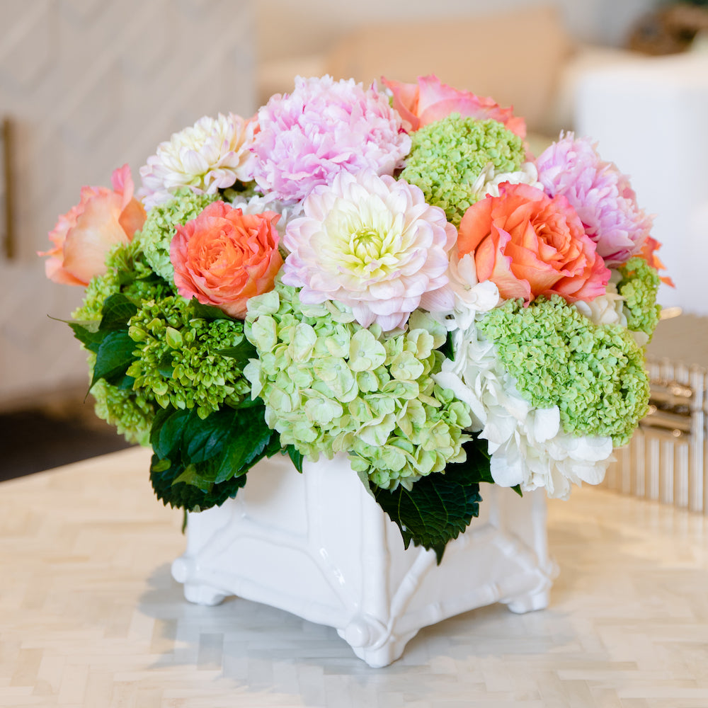 A white ceramic vase from Hive Floral Studio holds the Live Designer's Choice Floral Arrangement with green hydrangeas, pink dahlias, white flowers, and orange roses, displayed elegantly on a light-colored table.