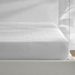 A neatly made bed features the Matouk Foundation Mattress Pad, an OEKO-TEX certified white quilted protector, in a modern bedroom with light walls and a wall-mounted lamp.
