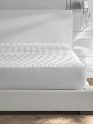 A neatly made bed features the Matouk Foundation Mattress Pad, an OEKO-TEX certified white quilted protector, in a modern bedroom with light walls and a wall-mounted lamp.