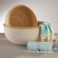 The Santo Rattan Bowl Collection by Zodax features two artisanal bowls with light wood and white finishes, displayed beside blue and white fringed cloths on a neutral surface.