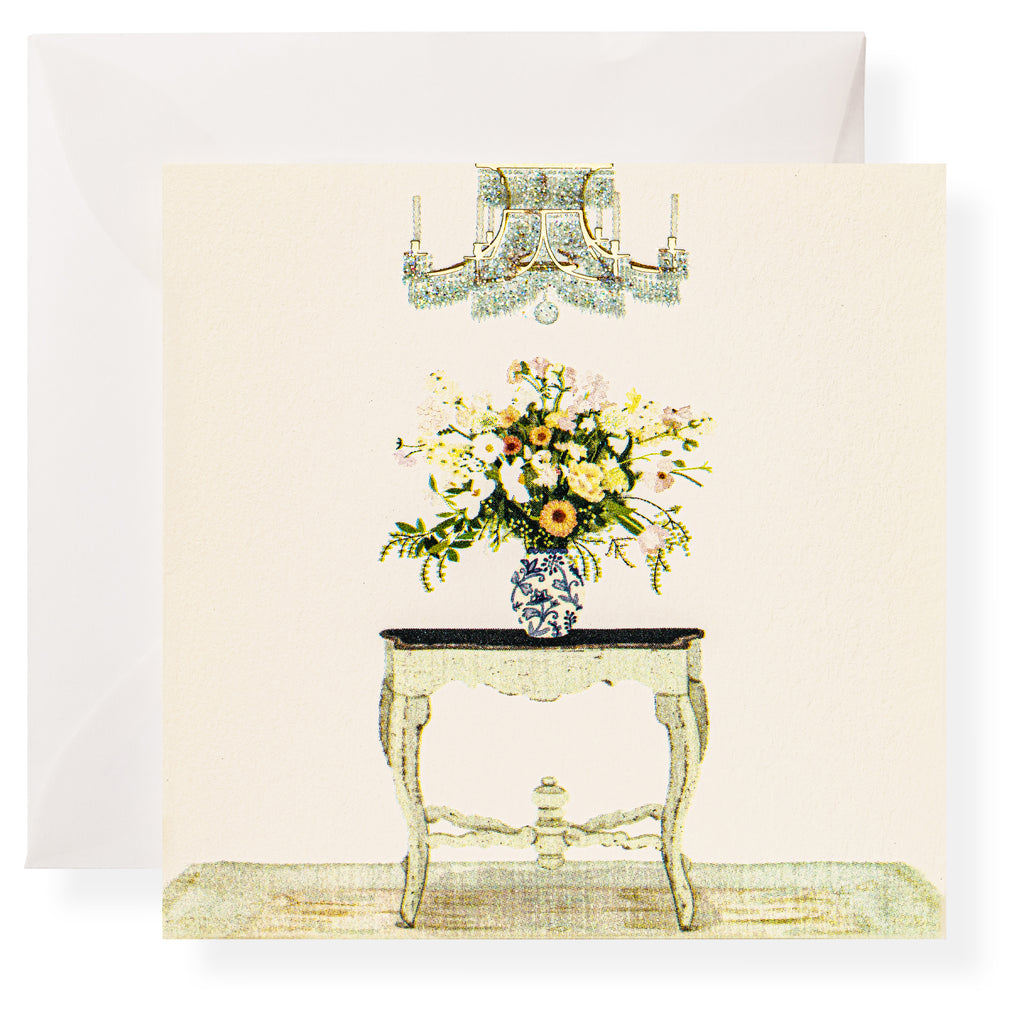A floral arrangement in a patterned vase sits on a small ornate table beneath a chandelier, with Karen Adams Dolce Vita Gift Enclosures and a white envelope in the background.