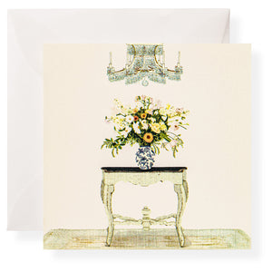 A floral arrangement in a patterned vase sits on a small ornate table beneath a chandelier, with Karen Adams Dolce Vita Gift Enclosures and a white envelope in the background.